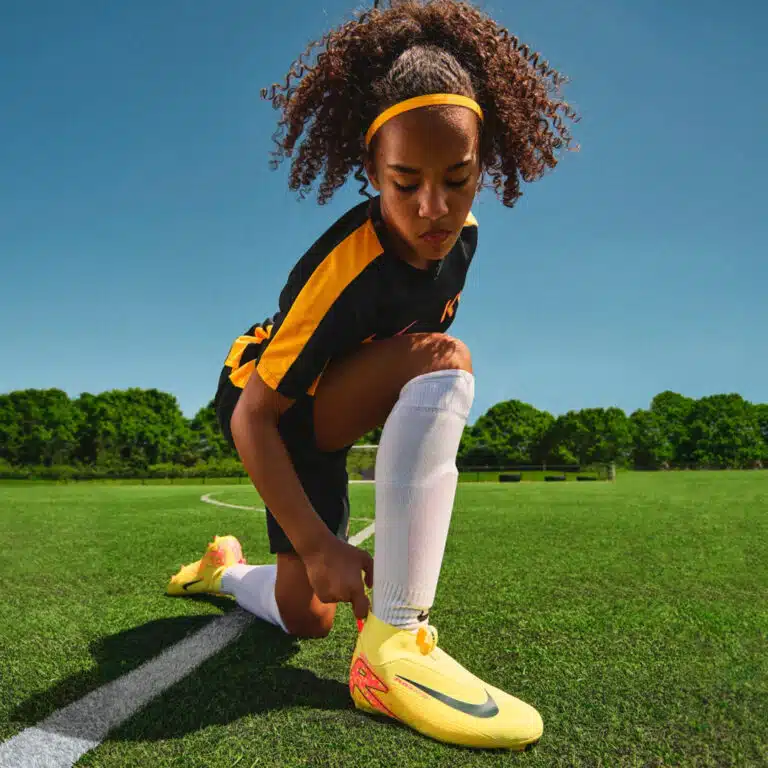 A girl lacing up yellow Nike Mercurial Superfly junior boots on a grass pitch